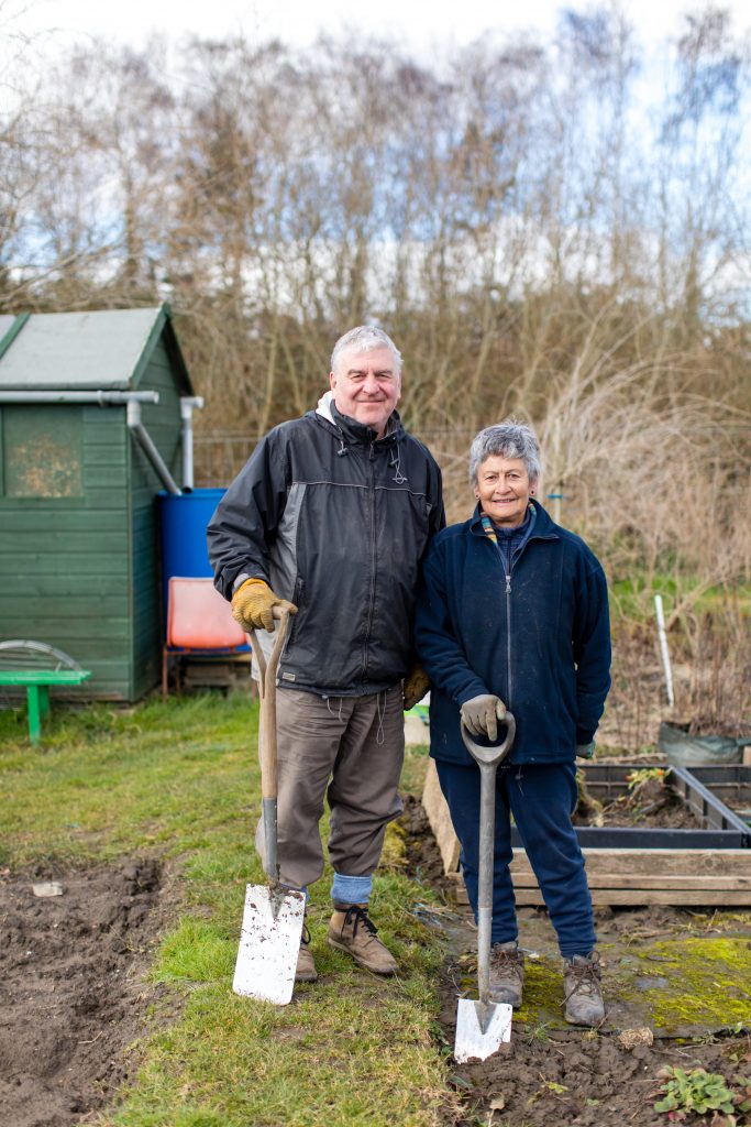 Older couple standing with their shovels in front of their allotment shed